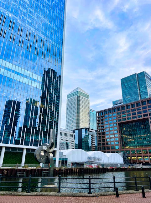 A cityscape featuring modern high-rise buildings with glass facades reflecting the sky and surrounding structures. The scene includes a waterfront with a black metal railing along the edge. One building has a prominent steel sculpture in front, while other buildings vary in height and design, including a distinctive white building with a rounded top. The sky is partly cloudy with patches of blue, and the overall environment appears clean and well-maintained, exemplifying urban architectural and structural elements in Canary Wharf. This image aligns with the professional cleaning and maintenance standards promoted by Carpet Cleaning SE8 for residential and commercial properties, emphasizing the importance of surface cleaning and sanitation in urban environments.