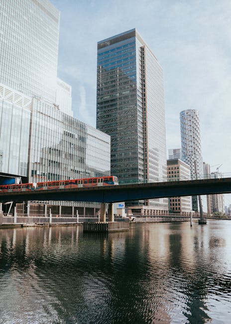 Image of a modern urban cityscape featuring several high-rise buildings with glass facades under a partly cloudy sky. A red train is visible crossing an elevated bridge over a river, reflecting the surrounding skyscrapers in the water. The scene depicts a bustling financial district with contemporary architectural styles, emphasizing clean, sleek surfaces and the reflective qualities of glass and metal. Bright natural lighting highlights the clarity and condition of the buildings and the water below, suggesting a well-maintained, vibrant area. This visual aligns with the services provided by Carpet Cleaning SE8 in surface cleaning, deep cleaning, and sanitisation within residential and commercial environments in the Canary Wharf E14 area.