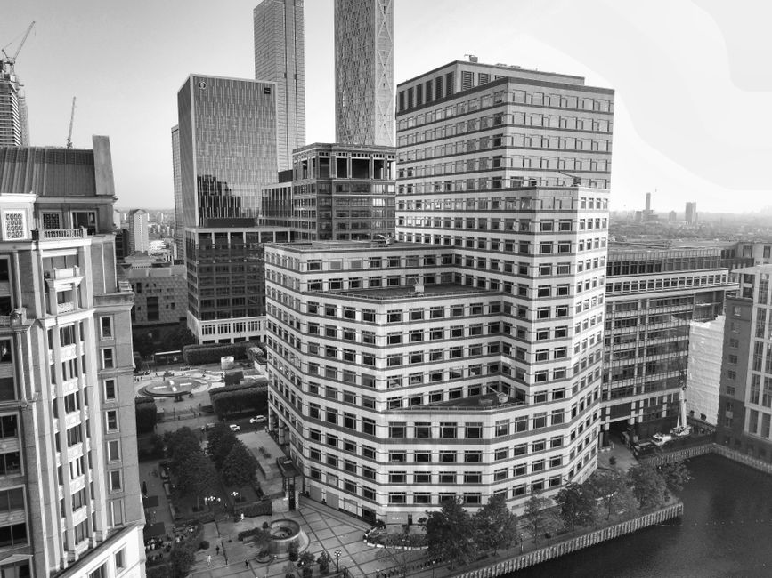 A cityscape featuring modern high-rise buildings with glass facades reflecting the sky and surrounding structures. The scene includes a waterfront with a black metal railing along the edge. One building has a prominent steel sculpture in front, while other buildings vary in height and design, including a distinctive white building with a rounded top. The sky is partly cloudy with patches of blue, and the overall environment appears clean and well-maintained, exemplifying urban architectural and structural elements in Canary Wharf. This image aligns with the professional cleaning and maintenance standards promoted by Carpet Cleaning SE8 for residential and commercial properties, emphasizing the importance of surface cleaning and sanitation in urban environments.