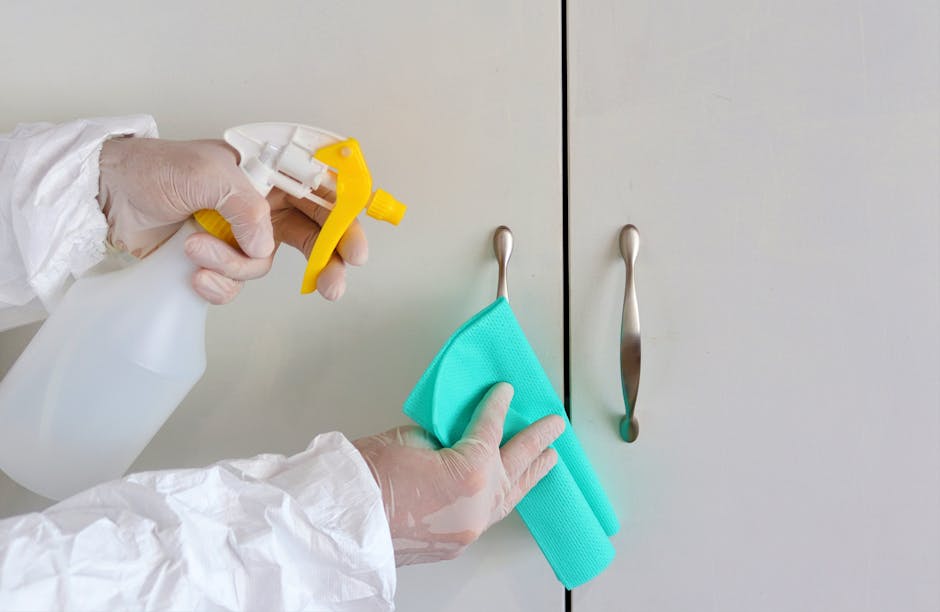 Close-up of a person wearing bright yellow rubber gloves, holding a yellow cleaning cloth. The scene takes place on a light gray countertop with a blue cleaning sponge placed nearby. The background is